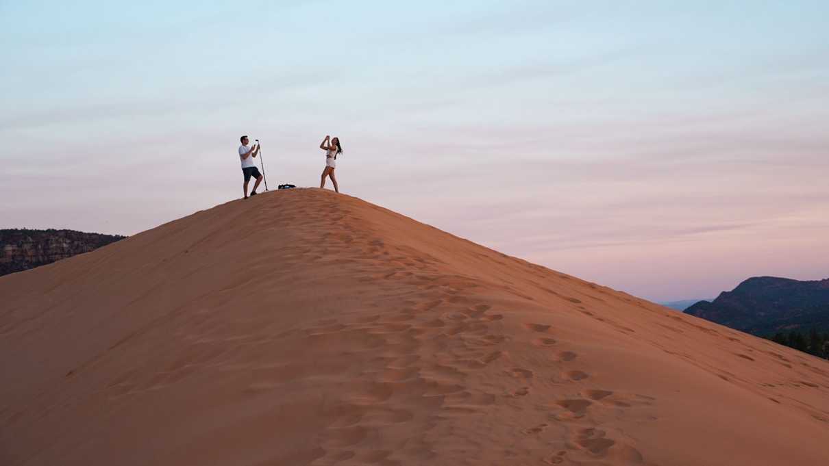 A photographer and model stand on top of sun lit sand dune